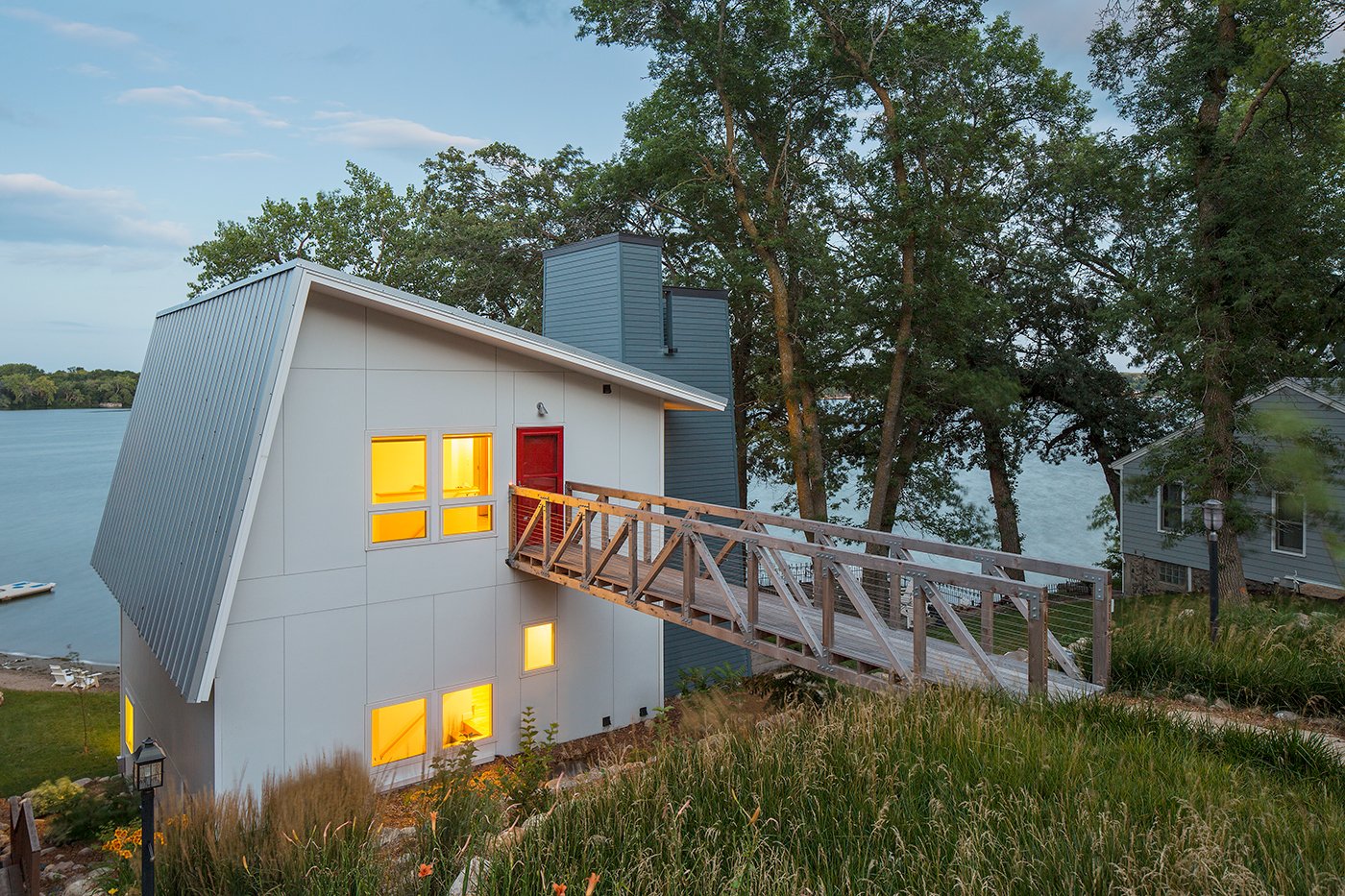 A modern lake home with standing-seam metal roof, white siding, and bright red front door.