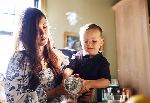 Inside a kitchen, a woman holds a young boy who is pouring water into a glass