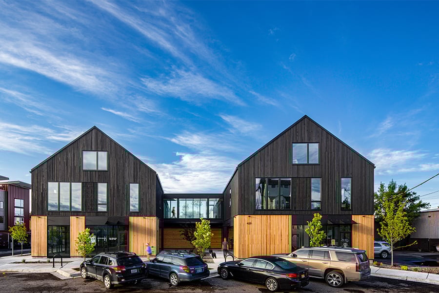 A street view of a mixed-use building with two parallel volumes connected via a skyway. 
