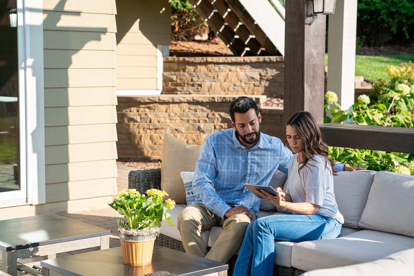 A man and woman sit on an outdoor sofa reading a tablet with hydrangeas and a green lawn in the background.