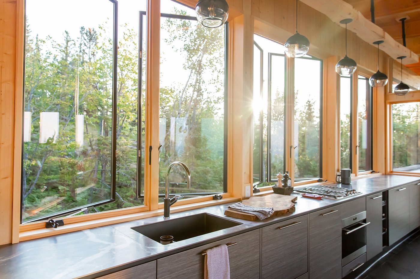 Light pours through a row of six open casement windows that serve as a backsplash in a kitchen with sleek modern cabinetry.