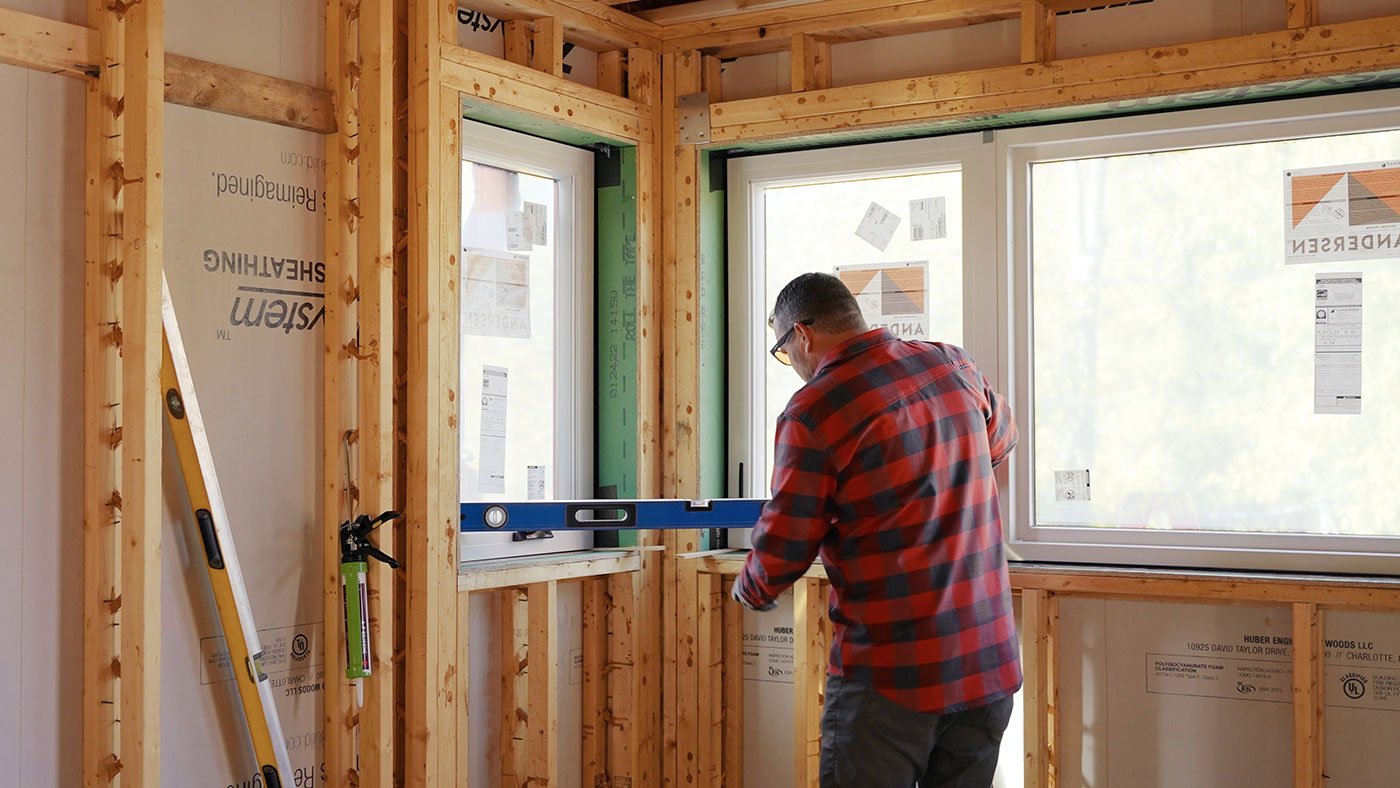 A builder stands inside a framed-out house and uses a level to check that a window is properly installed.