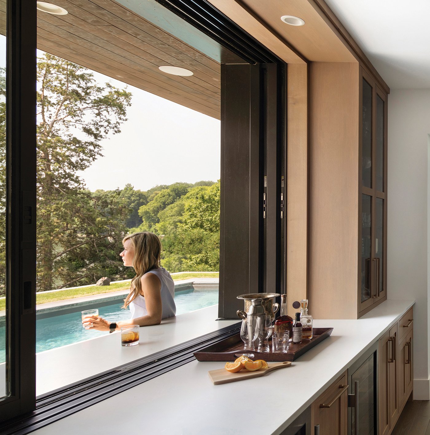 An open kitchen pass-through window sits flush with the countertop, which creates an outdoor bar where a woman sips her cocktail and looks out over the swimming pool.