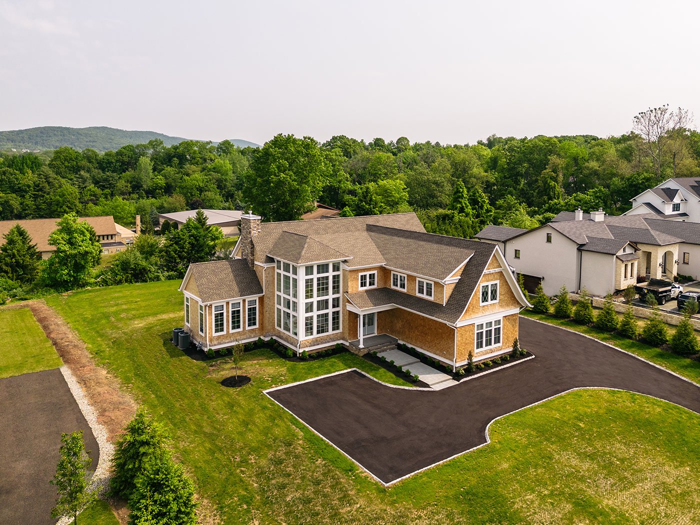 A home with brown wood shingles sits on a large plot of grassy land with a semi-circle driveway. The home has French doors and two stories of white windows with diamond and square window patterns
