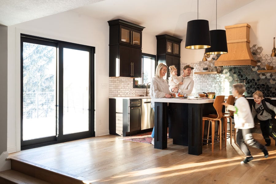 Parents laugh while their children race around a kitchen featuring black cabinetry and a black framed sliding glass door.