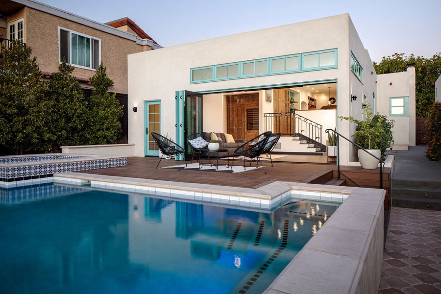 A white stucco home with aquamarine windows and doors has a bi-folding door that opens up the family room to a raised deck and backyard swimming pool.