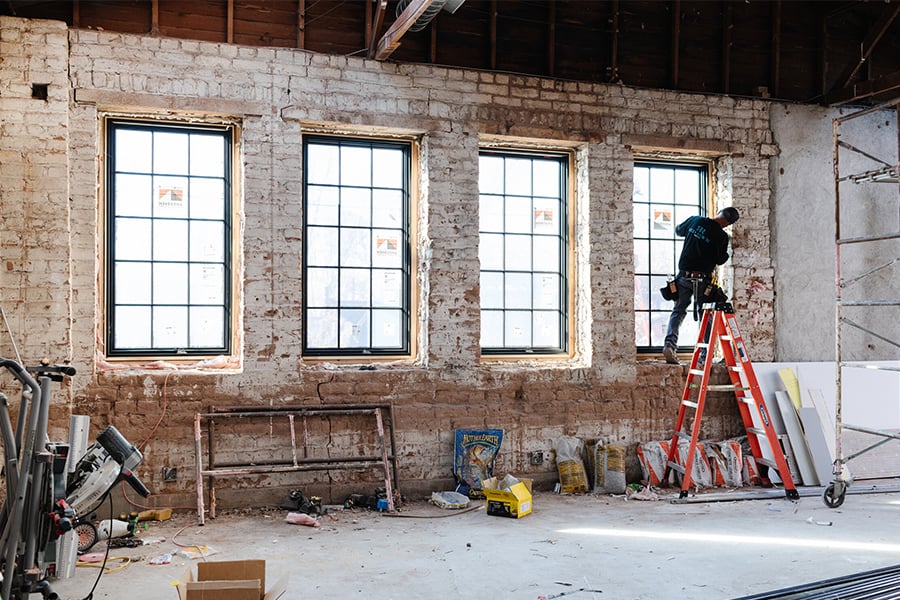 Inside a building with exposed brick walls, a man installs the last window in a row of four black-framed casement windows with grilles.