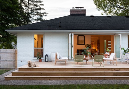 A dog lays on the deck outside an open bi-folding door built into the back of a ranch home.