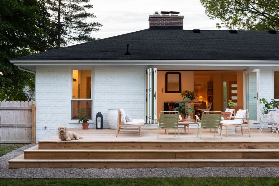 A dog lays on the deck outside an open bi-folding door built into the back of a ranch home.