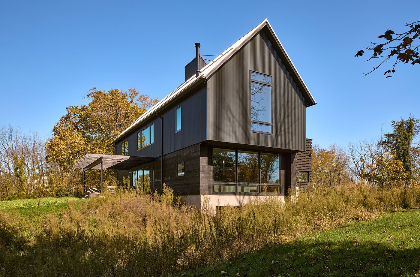 A barn-shaped home with monumental windows and a combination of black metal and shou sugi ban siding stands in a meadow.