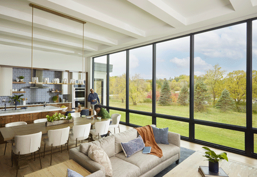 A couple enjoy their kitchen, which features a monumental wall of windows overlooking trees and a pond.