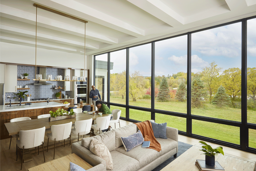 A couple enjoy their kitchen, which features a monumental wall of windows overlooking trees and a pond.